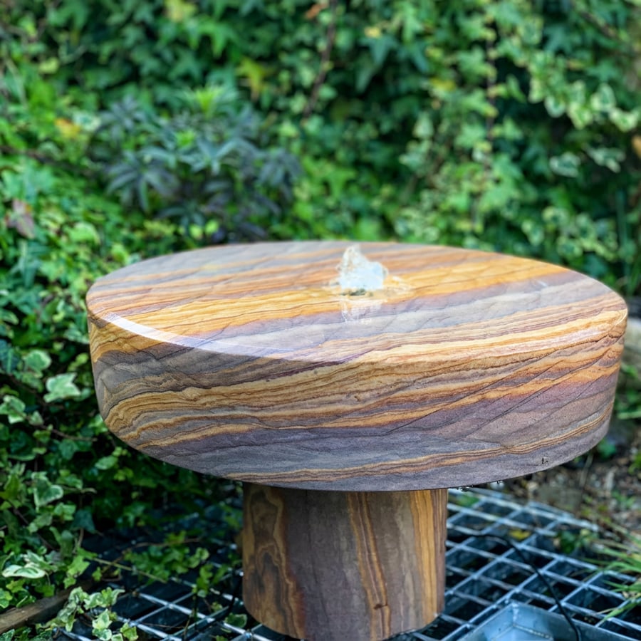 Rainbow sandstone millstone water feature at ground level in a UK wildlife garden with a sparrow drinking from the rim